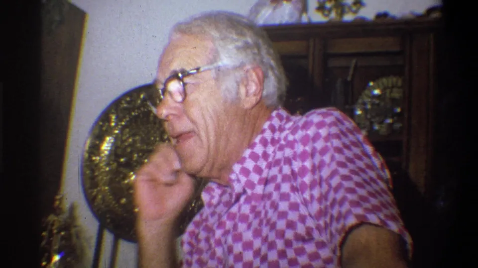 In 1973 Troy home-movie footage, the older man touches his chin while seated near wall decor.