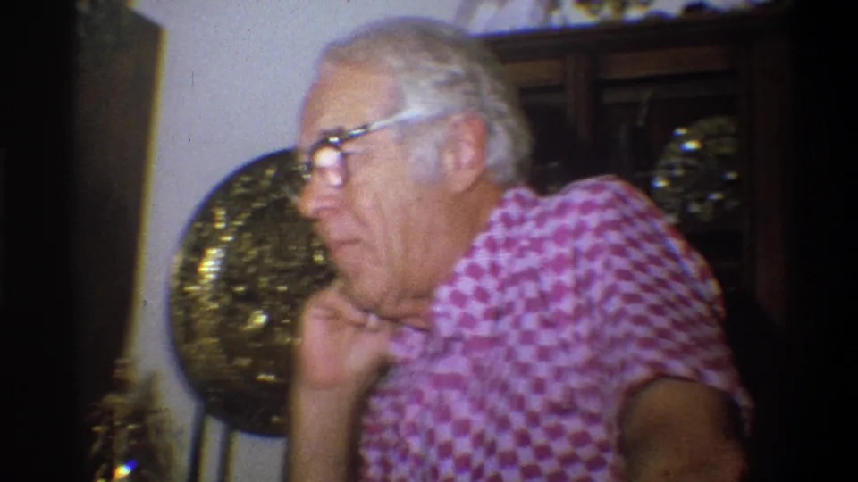 Older man wearing a pink checkered shirt rests his hand near his face in Troy, New York, 1973 home footage.