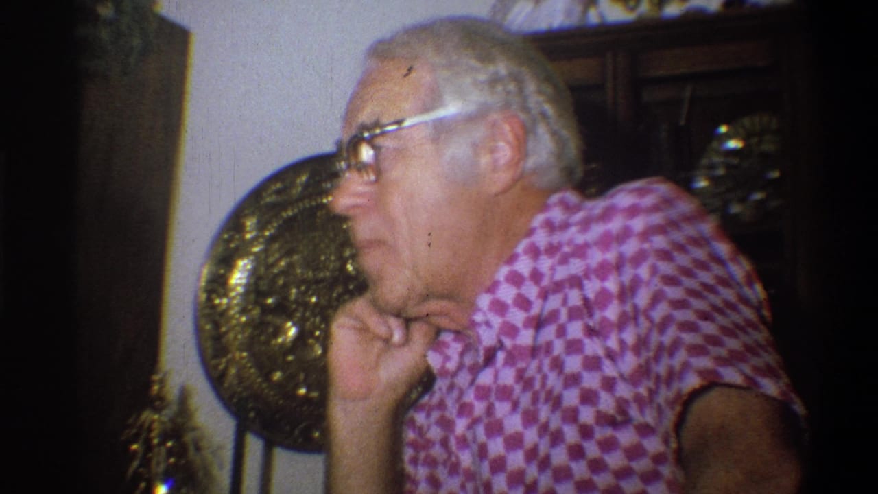 Older man in glasses and pink checkered shirt resting his hand near his chin indoors.