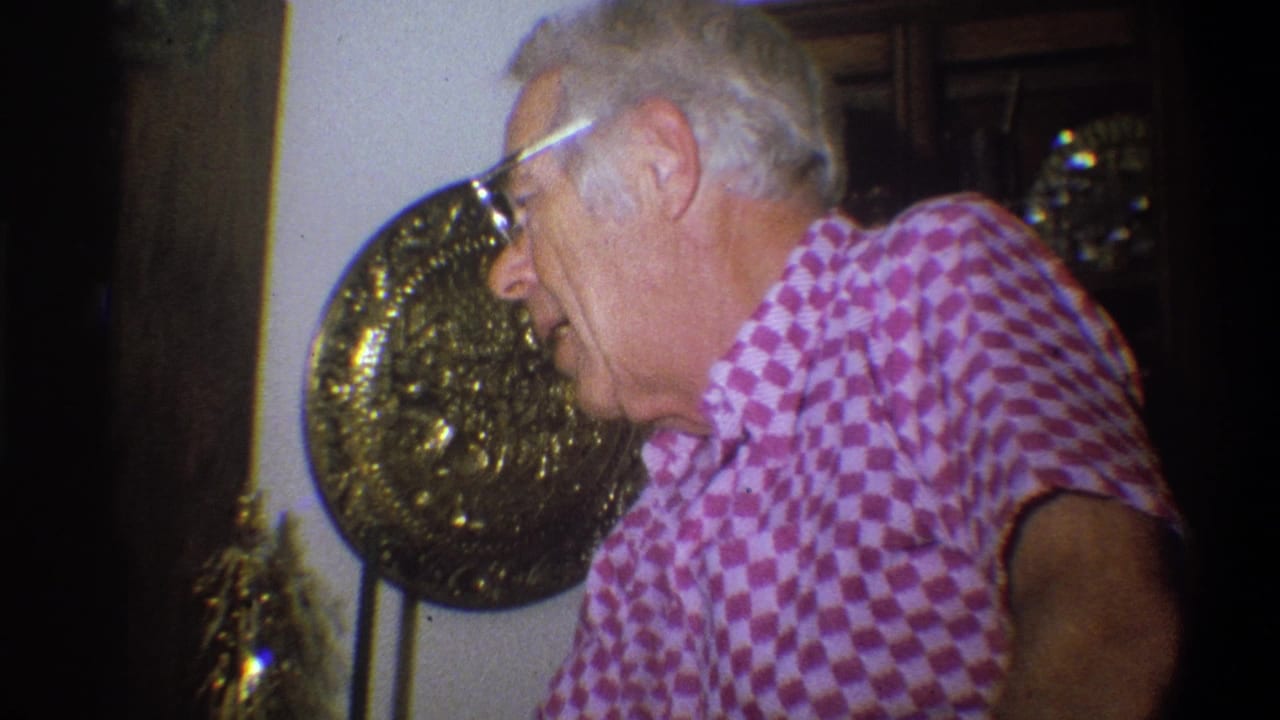 Profile view of an older man in glasses and a pink checkered shirt inside the Troy, New York family home setting.