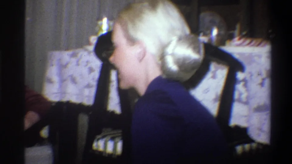 Blonde young woman with a bun and dark dress sits near a patterned tablecloth in a 1973 Troy home interior.