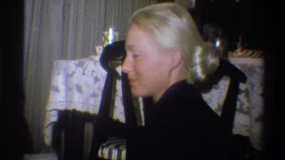 Blonde young woman in a dark dress sits in profile beside a dining table in the Troy, New York home-movie scene.