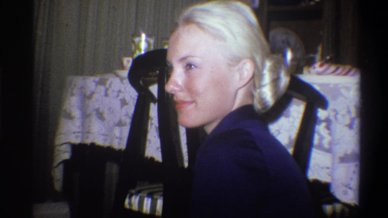 In a 1973 Troy, New York home-movie frame, a blonde woman sits indoors in a dark blue top beside a dining chair and tablecloth.