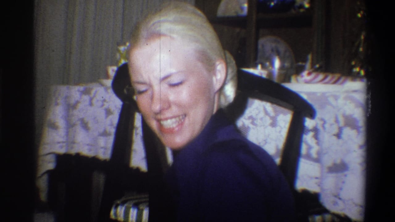 Blonde woman smiling with eyes lowered while seated near a table and chair.