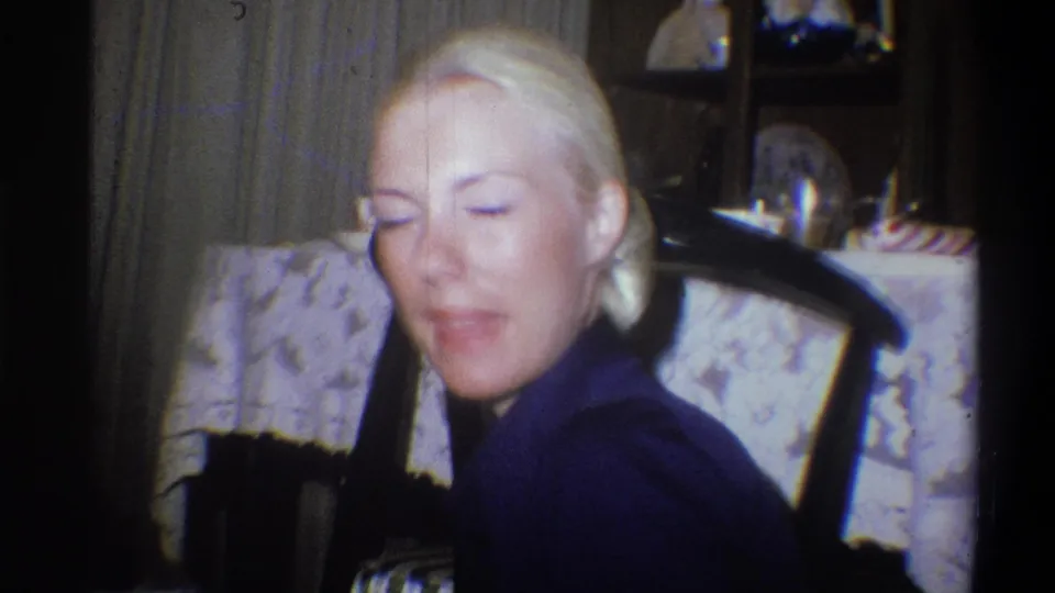 The close home-movie frame shows the blonde woman seated indoors with eyes closed, near a chair and patterned tablecloth.