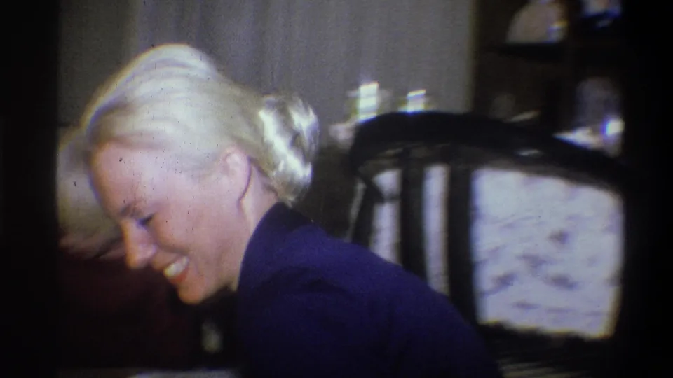 The woman laughs and leans forward in the 1973 Troy, New York home-movie footage, with a dining chair and table behind her.