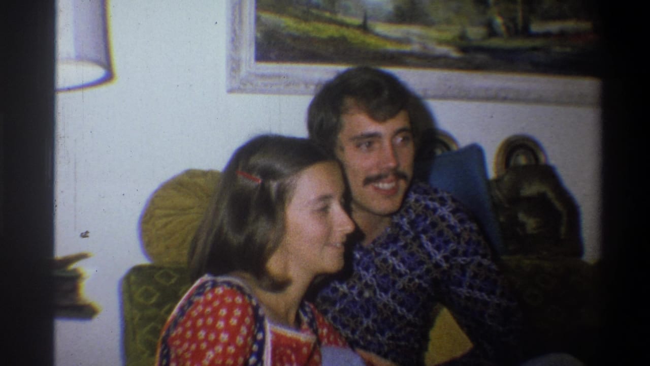 Woman in a red patterned dress sitting beside a mustached man in a blue patterned shirt.