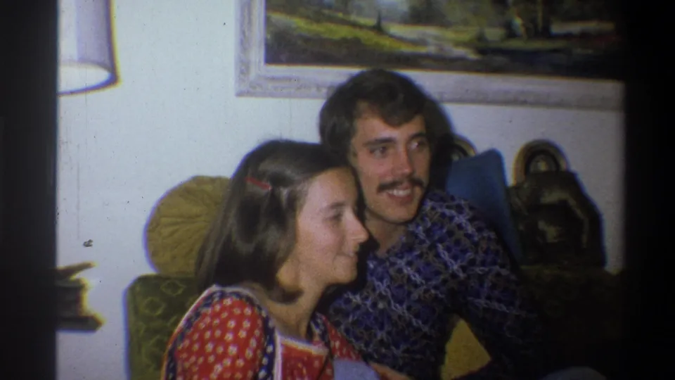 A woman and mustached man sit close together on a couch in a 1973 Troy, New York living room, with a framed landscape above them.