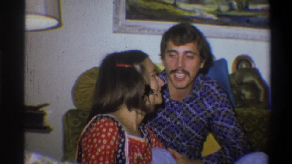 The woman turns toward the seated man in a 1973 Troy, New York home-movie view, with a lamp and wall art visible behind them.