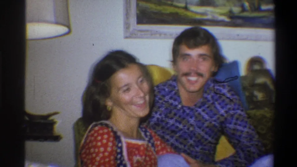 A smiling woman and mustached man sit together in a softly focused 1973 Troy, New York living-room home movie frame.