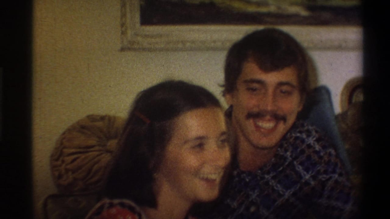 The couple smiles and looks toward someone off camera in a 1970s living room with framed art and cushions behind them.