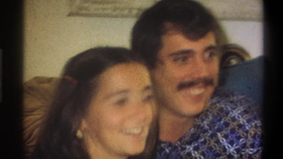 A close, softly focused 8mm frame shows the young woman and mustached man smiling together in the Troy home interior.