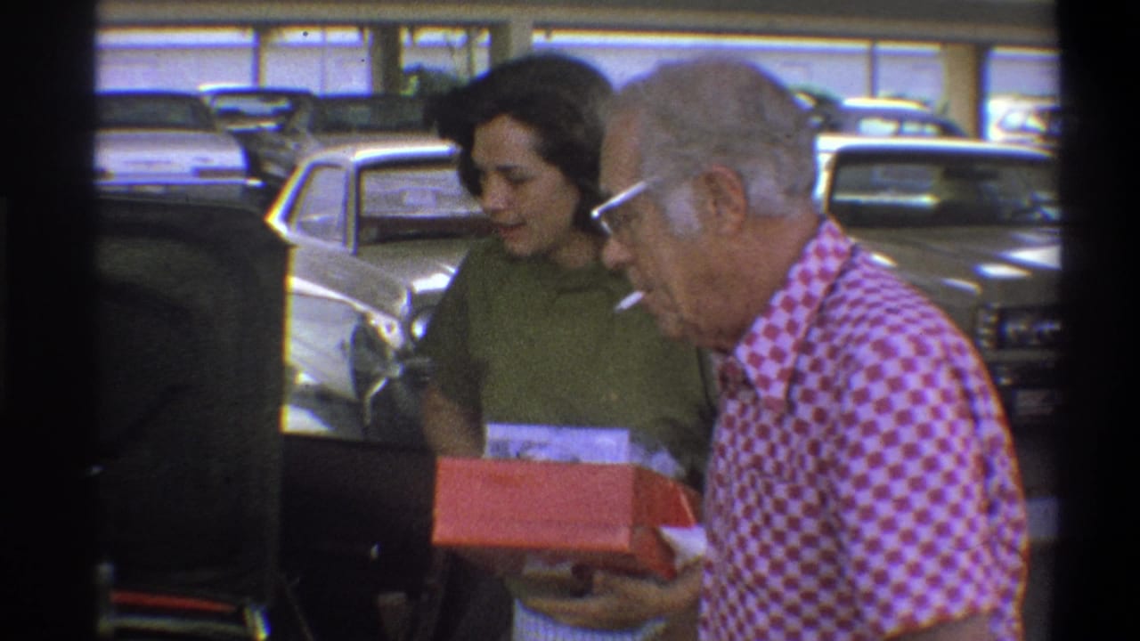 An older man in a red-and-white checkered shirt smokes while holding a red box near a woman in 1973 Troy footage.