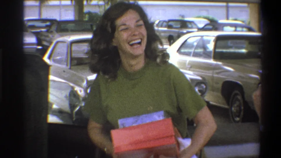 A woman laughs broadly while holding a red box in a Troy, New York parking garage with 1970s cars behind her.