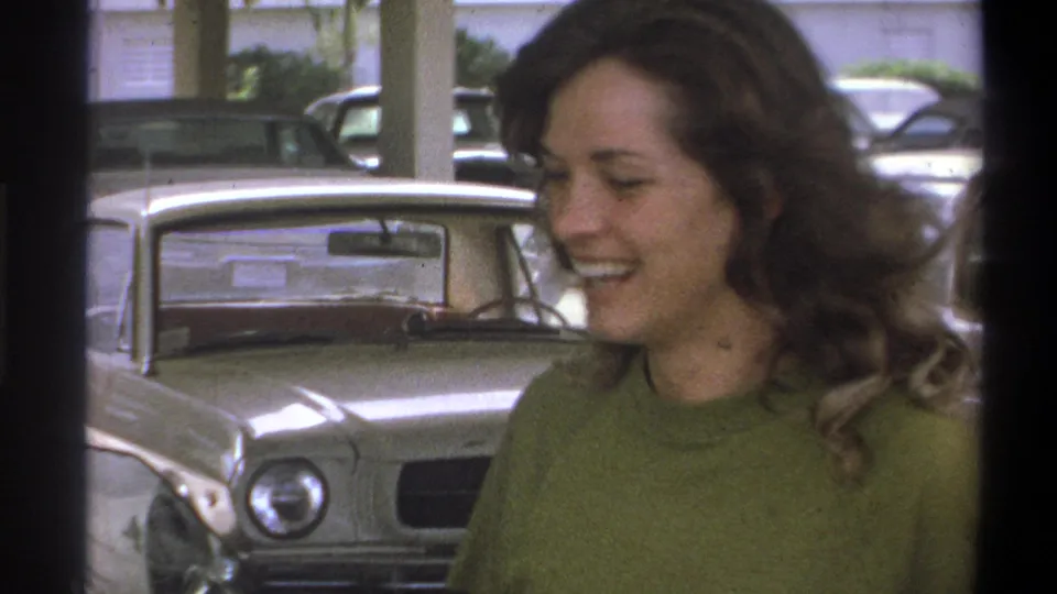 Close 1973 Troy home-movie view of a smiling woman near a light-colored sedan inside a parking garage.