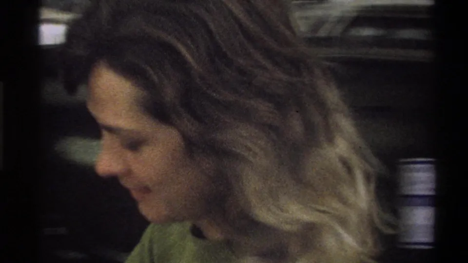 A young woman in profile appears beside a dark parked car in the 1973 Troy, New York home-movie scene.