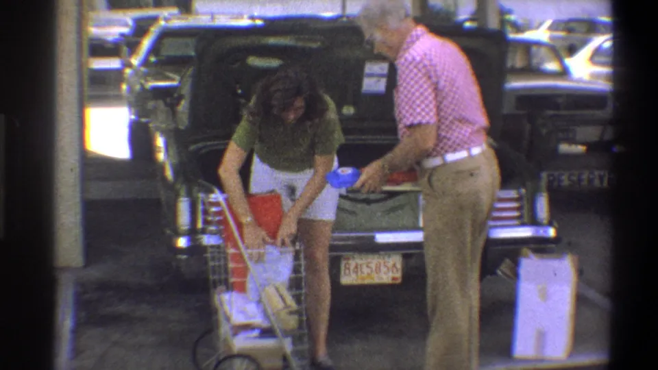 A young woman bends over groceries in a cart while an older man stands beside an open car trunk in Troy, New York, 1973.