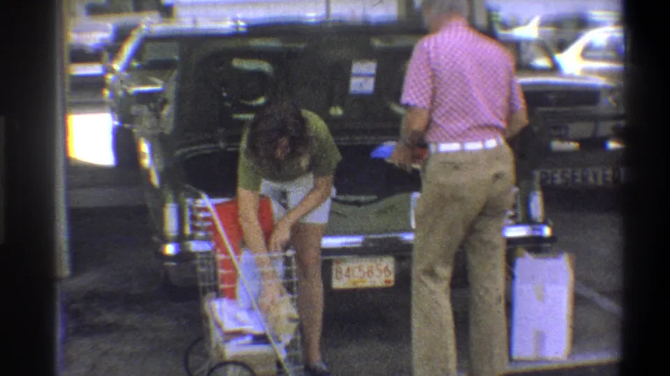 The older man faces the open trunk as the young woman continues handling grocery bags from the cart in the 1973 Troy scene.