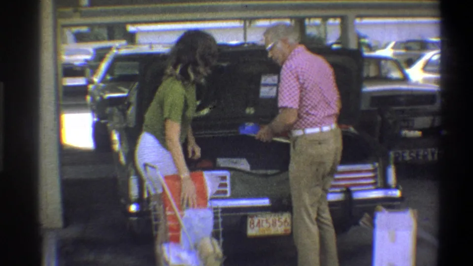 Man and woman facing each other beside a dark American sedan in a parking lot with shopping cart, Reserved parking sign visible, Troy, New York, circa 1973.