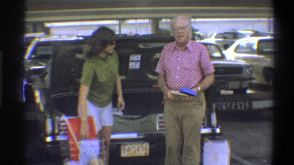 Older man in pink checkered shirt and woman in green top wearing sunglasses face toward camera in a parking lot, Troy, New York, circa 1973, faces identifiable.