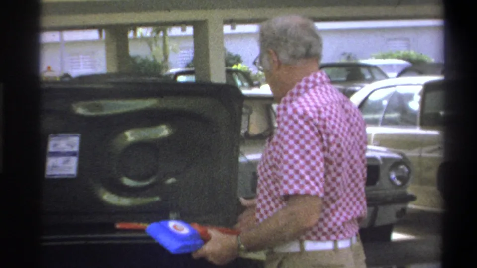 The man holds a blue object near the open trunk as the home movie records travel preparation in Troy, New York.