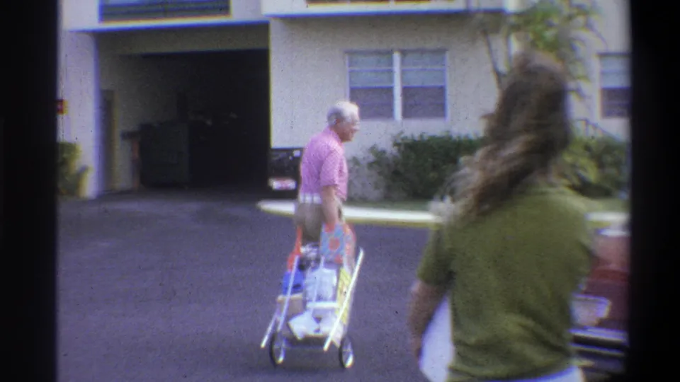 Two adults move away from the car with a small luggage cart outside a parking structure in 1973 Troy footage.