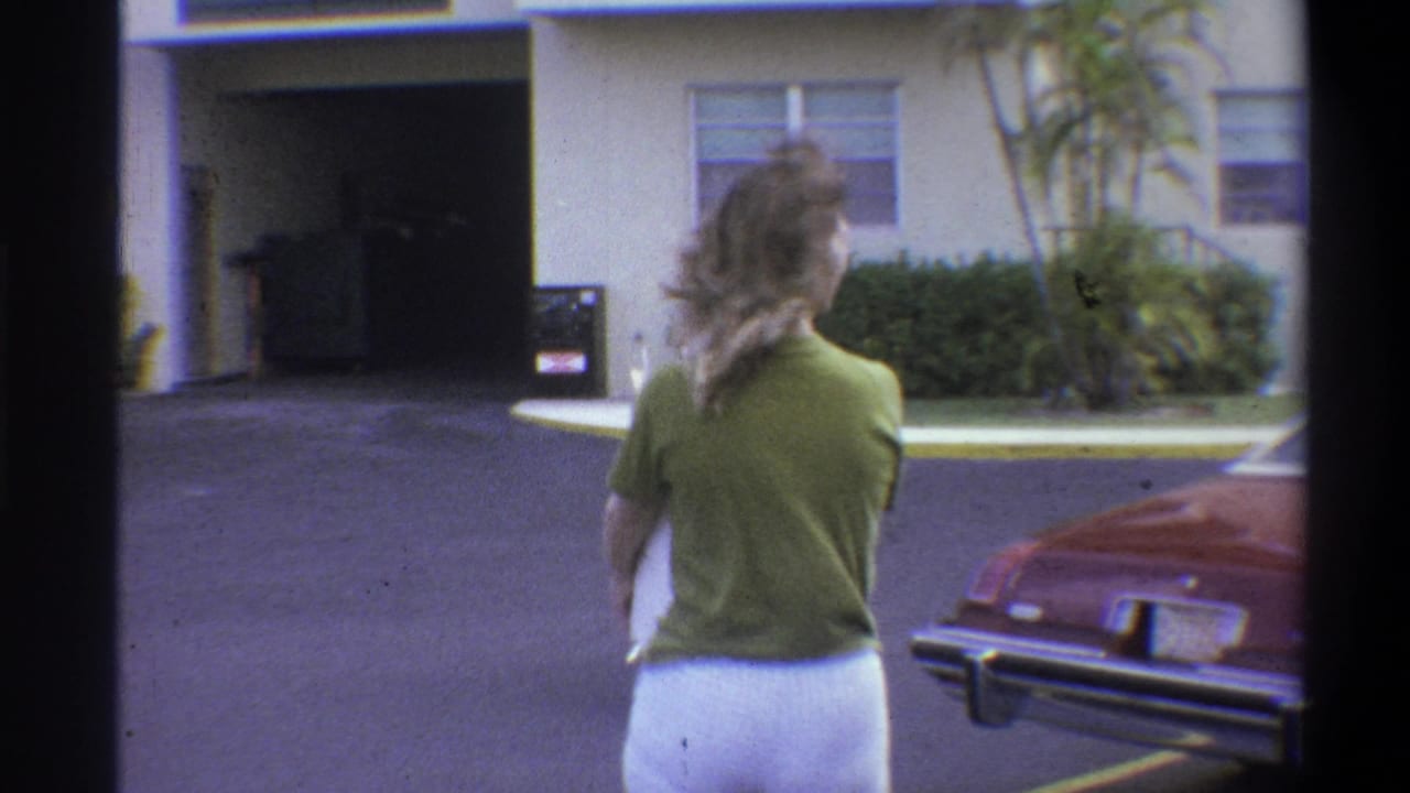 Woman in green top and white shorts walks away from camera in a residential complex parking area, circa 1973; dark-red classic American car and large palm trees visible.