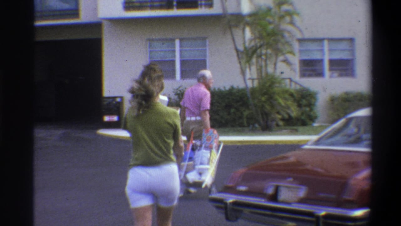 Couple walking away from camera — woman in green top and man in pink shirt pushing a colorful stroller — with a dark-red American car to the right.