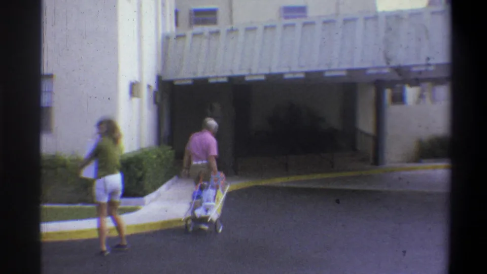 Couple with stroller walking toward the covered entrance of a mid-century apartment building in what appears to be a warm-climate residential setting, circa 1973.