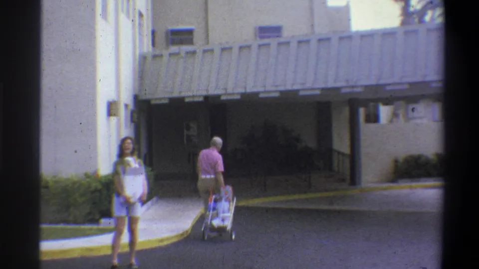 Couple with stroller continuing past the covered entrance of a residential apartment building, seen from behind, circa 1973; building canopy and pale facade clearly visible.