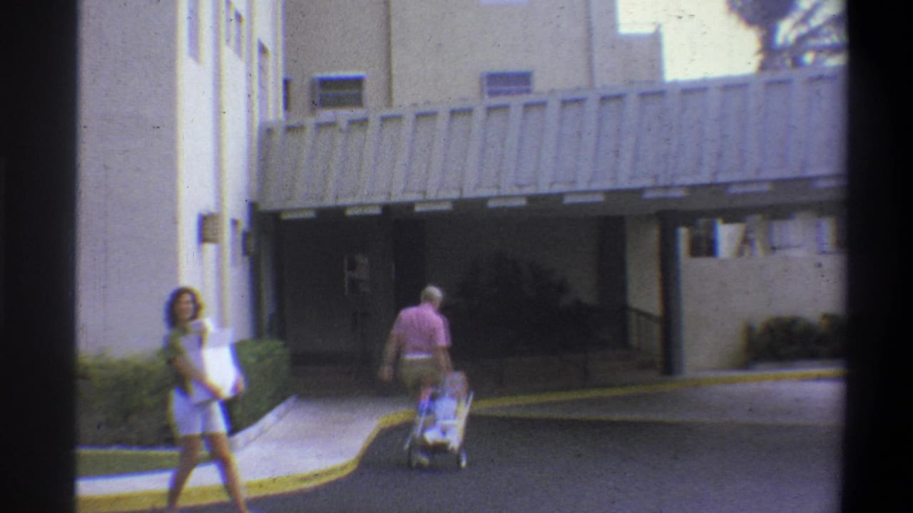 1973 8mm home movie frame showing a woman in teal top and shorts carrying a box and an older man in pink shirt pushing a luggage cart toward a modern building entrance, Troy, New York.