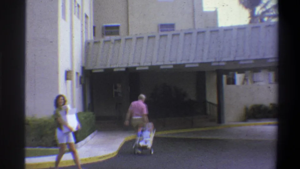 1973 8mm home movie frame showing a woman in teal top and shorts carrying a box and an older man in pink shirt pushing a luggage cart toward a modern building entrance, Troy, New York.