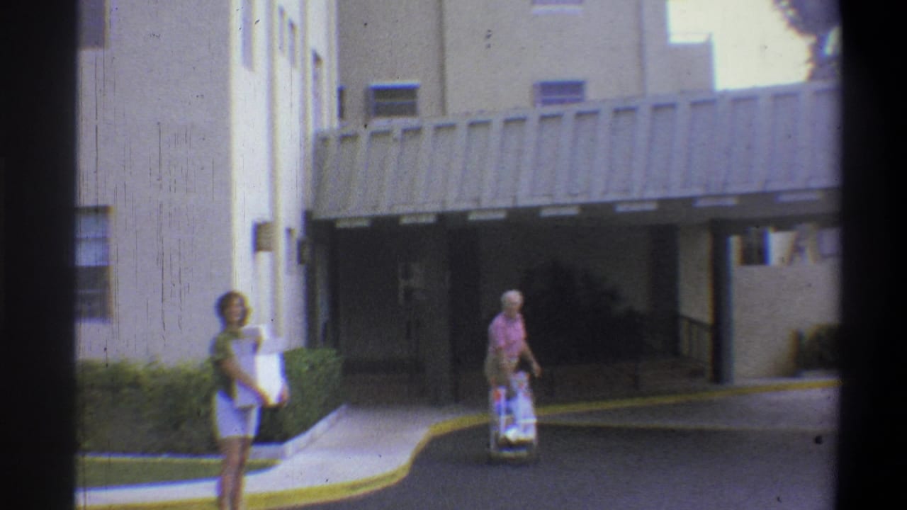 Two figures, a woman with a box and an older man pushing a cart, walking toward a covered building entrance in natural daylight.