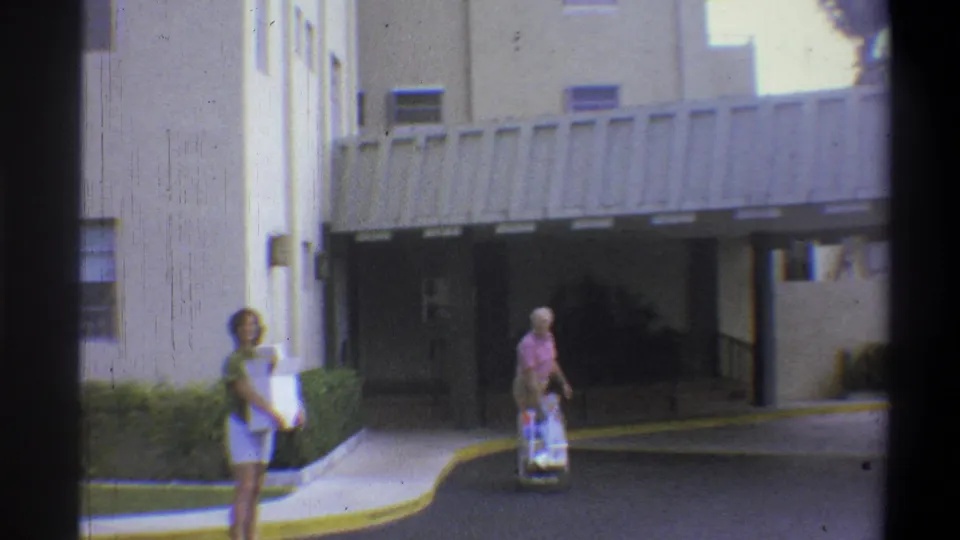 1973 8mm frame showing a woman and an older man with a luggage cart approaching a modern building entrance; similar wide angle to the opening frame, Troy, New York.