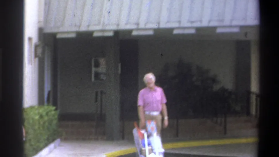 Close 8mm frame from 1973 showing an older man with white hair in a pink sport shirt pushing a wheeled luggage cart toward glass entrance doors, likely Troy, New York.