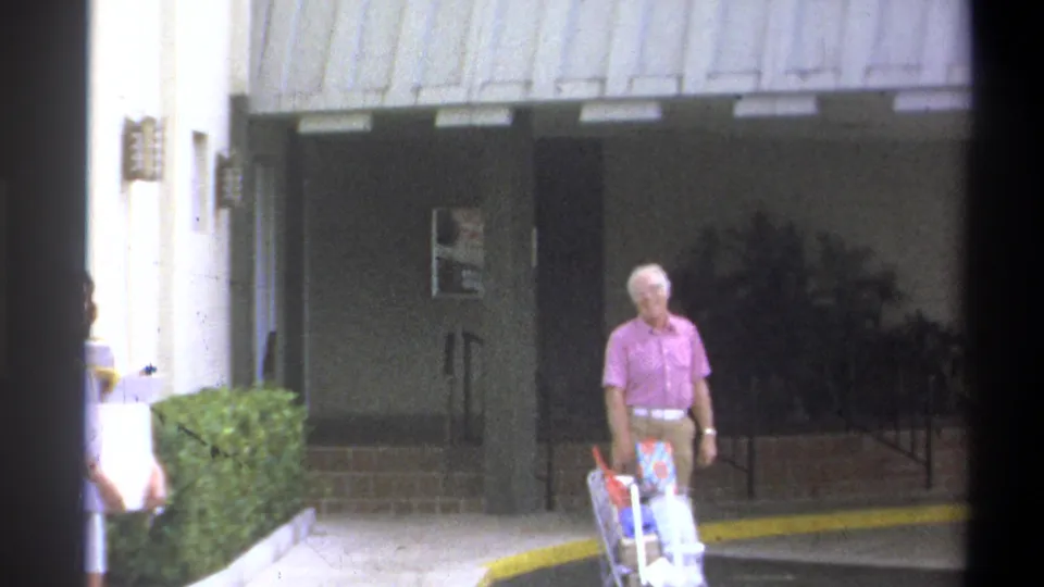 Final 8mm frame from 1973 clip showing older man in pink shirt with luggage cart at a modern building entrance, Troy, New York; second figure partially visible at left.