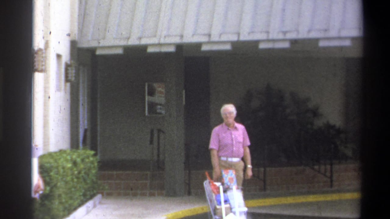 Older man in pink short-sleeve shirt pushes a laden shopping cart along a concrete path outside a covered building entrance, Troy, New York, 1973.