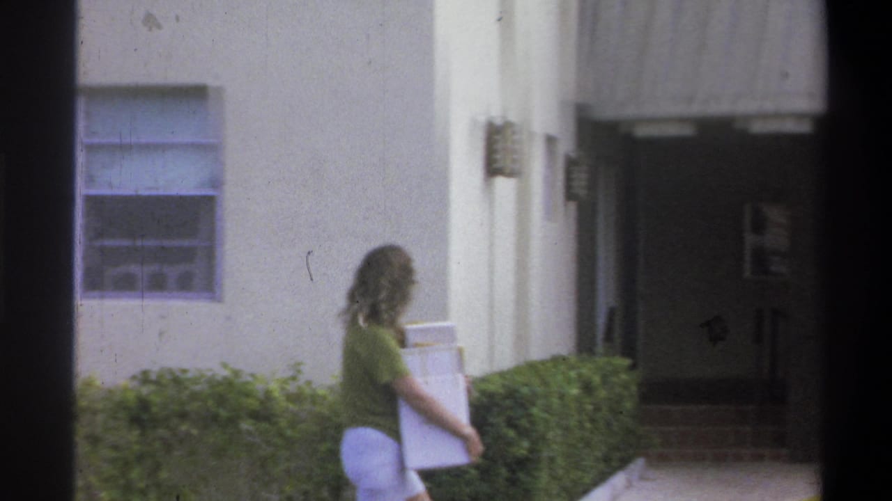 Woman in green shirt carrying a large white flat object along a concrete path with trimmed hedges beside a light-colored building.