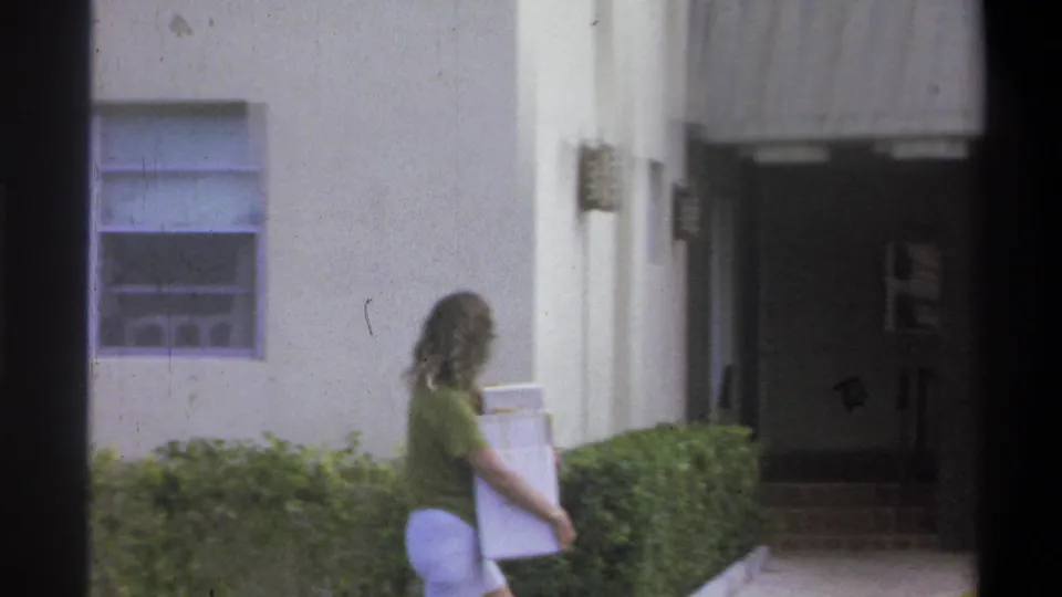 Woman in green short-sleeve shirt and blue trousers carries a large white box or panel beside well-trimmed hedges outside a building, Troy, New York, 1973.