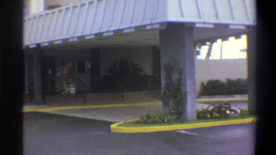 Establishing wide shot of a mid-century covered entrance canopy on concrete pillars with a yellow curb island, small figures with cart visible in shade, Troy, New York, 1973.