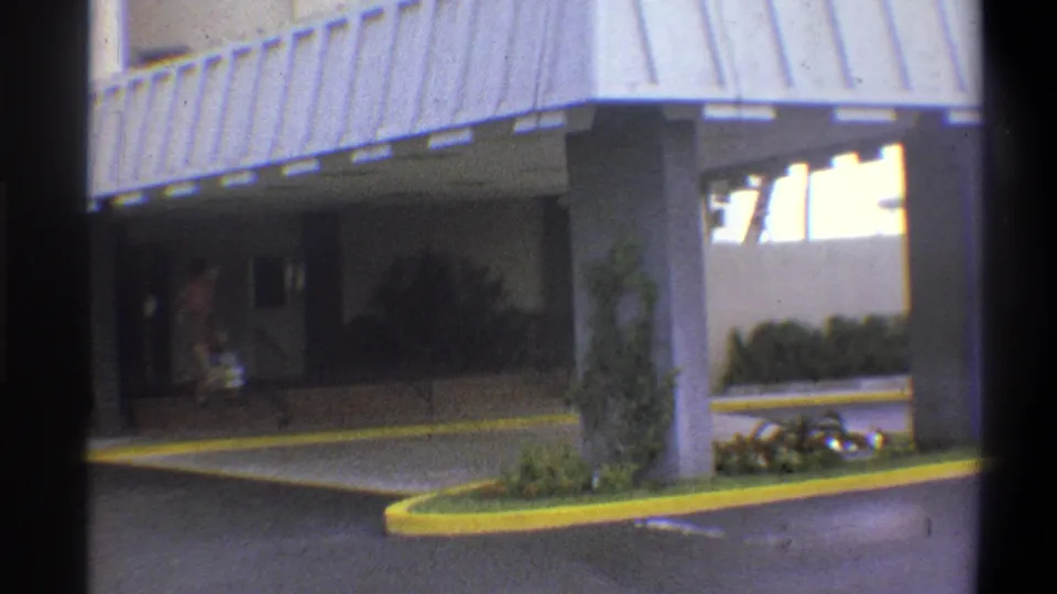 Continuation wide shot of the covered entrance canopy at a mid-century building, figures partially visible carrying items beneath it, yellow curb island prominent, Troy, New York, 1973.