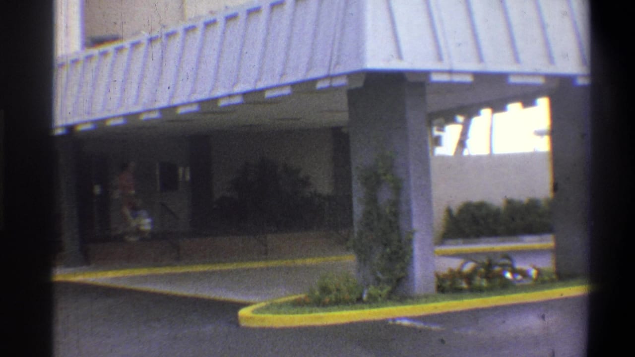 Hotel entrance with corrugated metal canopy and yellow-painted curved driveway curb; adult figure partially visible under awning; 8mm home movie, claimed 1973 Troy, New York.