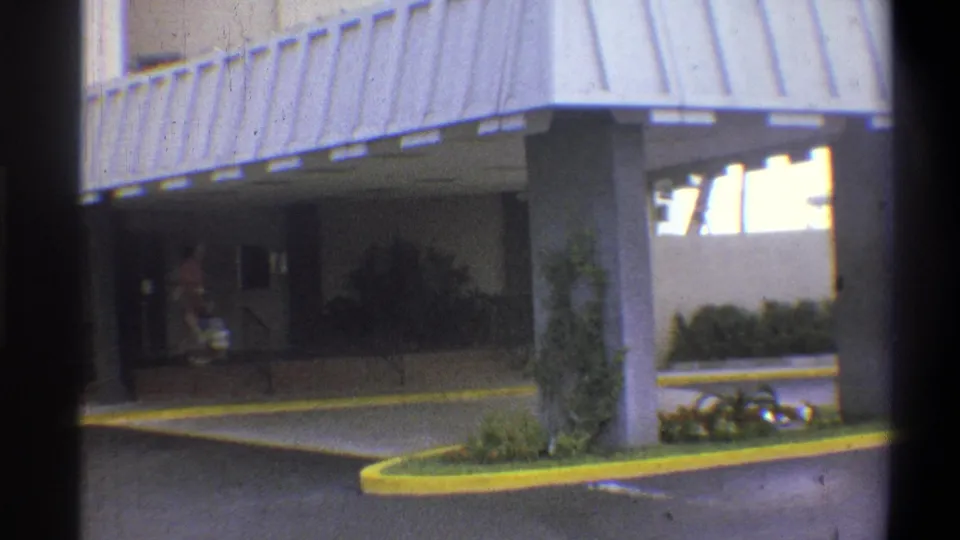 Hotel entrance with corrugated metal canopy and yellow-painted curved driveway curb; adult figure partially visible under awning; 8mm home movie, claimed 1973 Troy, New York.