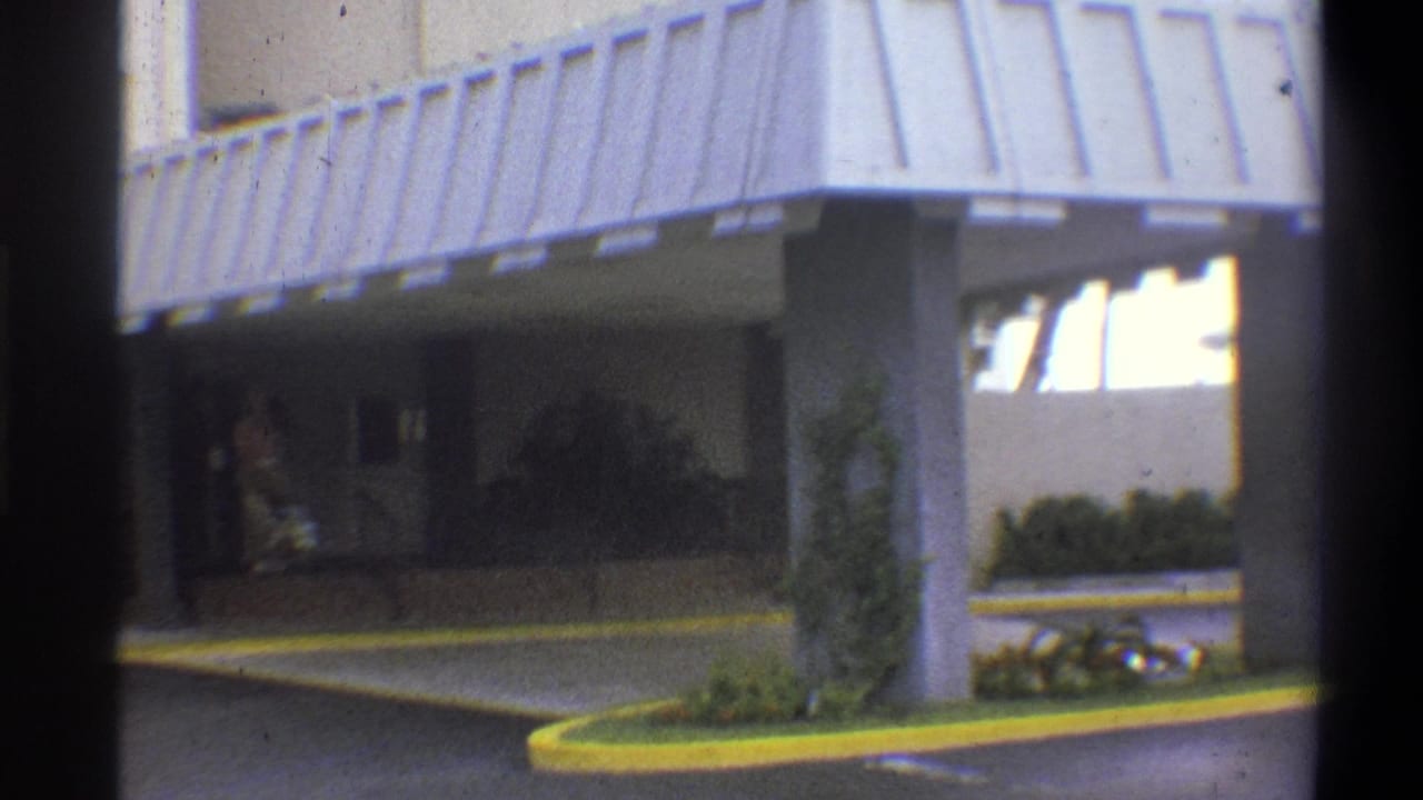 Hotel forecourt with corrugated metal awning and yellow-painted curved driveway; figure partially visible at right; 8mm home movie, claimed 1973 Troy, New York.