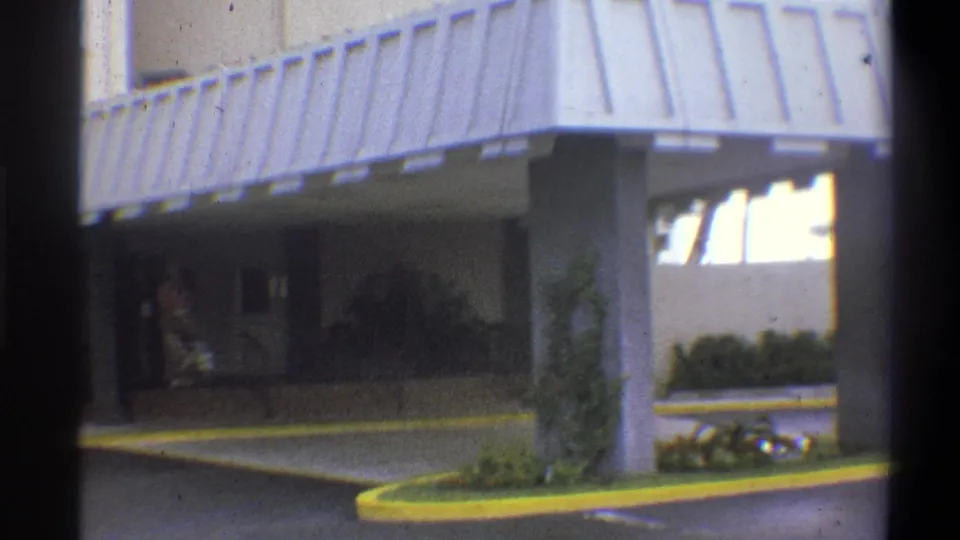 Hotel forecourt with corrugated metal awning and yellow-painted curved driveway; figure partially visible at right; 8mm home movie, claimed 1973 Troy, New York.