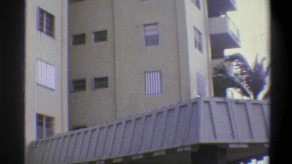 Multi-story beige hotel exterior with window louvers and corrugated awning at base; palm fronds visible at right; 8mm film, claimed 1973 Troy, New York.