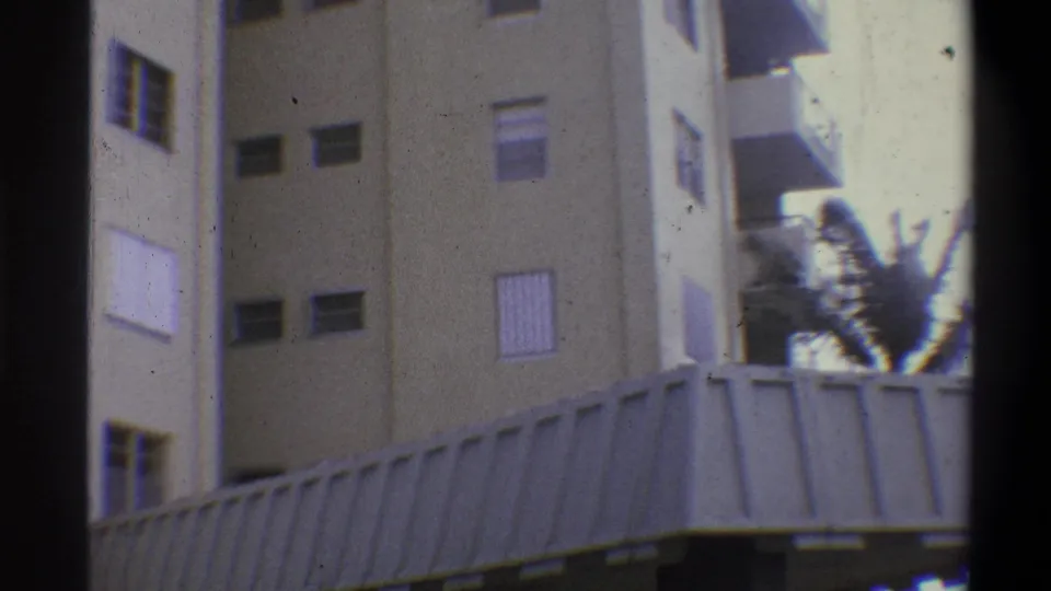 Hotel upper facade with palm fronds in motion at right; multi-story beige building with window louvers; 8mm home movie, claimed 1973 Troy, New York.