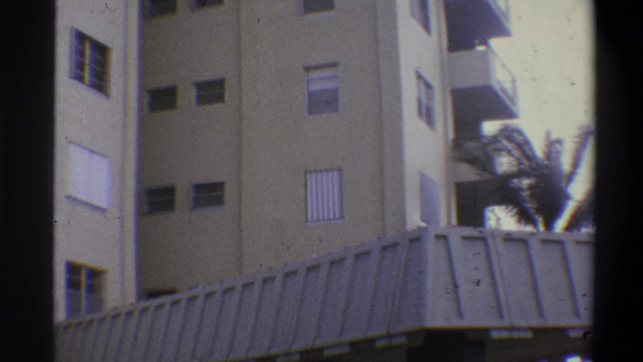 Handheld 8mm low-angle view of a tall tan concrete building with louvered shutters and balconies; a palm tree sways in strong wind at upper right, circa 1973.