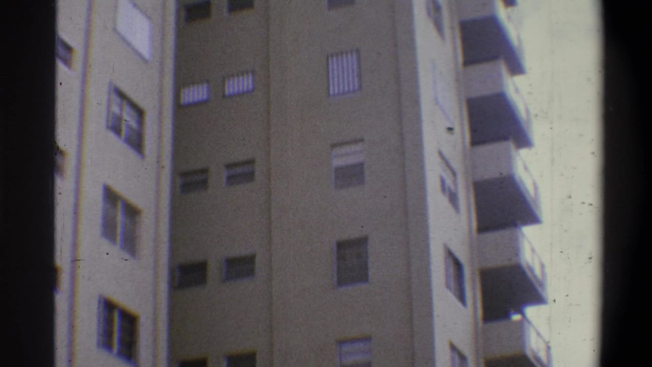 Handheld 8mm upward view of a tall tan concrete building with rows of louvered shutters and projecting balconies; palm tree no longer in frame, circa 1973.