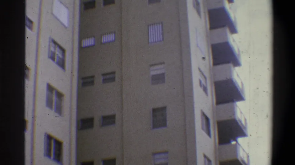 Handheld 8mm upward view of a tall tan concrete building with rows of louvered shutters and projecting balconies; palm tree no longer in frame, circa 1973.